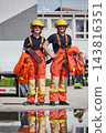 Two Firefighters in Orange Protective Gear and Yellow Helmets Standing With Reflective Puddle Image 143816351