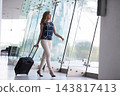 Businesswoman walking through modern airport terminal pulling rolling suitcase and looking out large windows 143817413