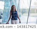 Young professional woman walking through modern airport terminal with rolling suitcase and smartphone 143817425