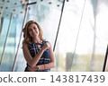 Confident young businesswoman smiling in modern office hallway by large glass windows, casual professional portrait 143817439