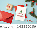 Merry christmas text on white card with red envelop, gift box, red ribbon and pine branch xmas ornament on blue background 143819169
