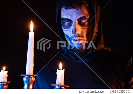 Candlelit portrait of hooded person with skull face paint holding lit candles under dramatic blue and orange light for Halloween 143820062