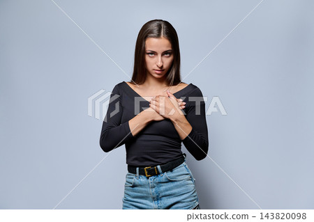 Serious young woman holding hands on her chest on gray background Serious young woman holding hands on her chest on gray background 143820098