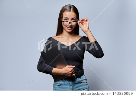 Young woman holding closed laptop and adjusting glasses in studio shot Young woman holding closed laptop and adjusting glasses in studio shot 143820099