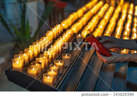 Person holding a smartphone near rows of lit votive candles inside a historic church in France, capturing a moment of quiet reflection and prayer 143820390