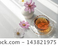 玻璃杯裡的茶和粉紅色的波斯菊,在秋天的陽光下閃閃發光,映襯著白色背景——完美的下午茶場景。 玻璃杯裡的茶和粉紅色的波斯菊,在秋天的陽光下閃閃發光,映襯著白色背景——完美的下午茶場景。 143821975