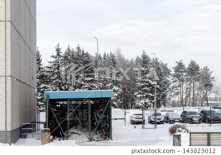 Bicycle storage shelter, covered bike rack parking lot near apartment building. Bikes parked under snowy pine trees in residential courtyard. Cars in winter parking lot, cloudy urban 143823012
