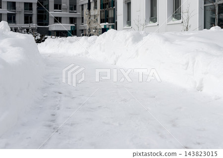 Snowdrift banks line snow covered path, shoveled walkway cleared for winter walk. Footprints and trail on snowy pile near apartment buildings. Urban residential district, overcast cityscape. Snowdrift banks line snow covered path, shoveled walkway cleared for winter walk. Footprints and trail on snowy pile near apartment buildings. Urban residential district, overcast cityscape. 143823015