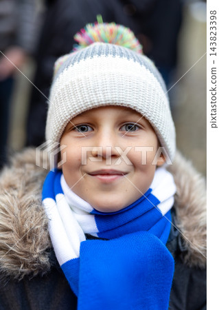 Portrait cute little child boy in winter hat blue scarf looks camera gentle smile. Outdoor street closeup view kid innocence warmth and cheerful mood in cold season with soft bokeh 143823398