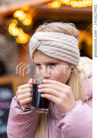 Teenage girl drinks warm kinderpunsch at Christmas market, wrapped in pink jacket and knitted headband. Cozy winter portrait with festive lights captures seasonal warmth, holiday mood 143823402