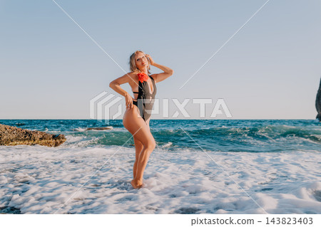 Coastal vacation getaway featuring mature woman posing gracefully in black swimwear by ocean surf during sunset 143823403