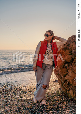 Woman beach sunset model in red puffer vest and sunglasses leaning on a rock at a pebble shore during golden hour Woman beach sunset model in red puffer vest and sunglasses leaning on a rock at a pebble shore during golden hour 143823476