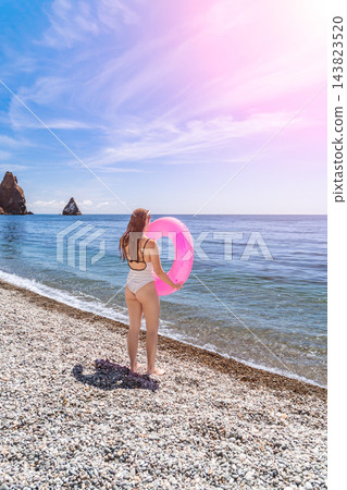 Woman beach swimsuit standing on pebble shore with pink inflatable ring enjoying summer vacation by blue ocean waters under bright sun 143823520