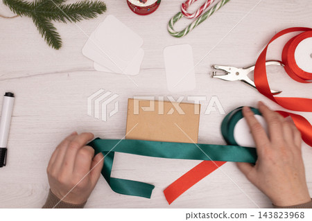 Hands wrap a kraft gift box with green ribbon at a festive Christmas gift station Hands wrap a kraft gift box with green ribbon at a festive Christmas gift station 143823968