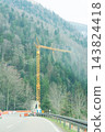 Construction equipment stands tall on a mountain road, surrounded by towering trees and steep slopes under a hazy sky at the edge of spring 143824418