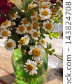 A beautiful bouquet of small white chamomile flowers with golden yellow centers arranged in a translucent green glass vase on a wooden table. Close-up floral photography, rustic home decor  143824708