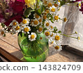A beautiful bouquet of small white chamomile flowers with golden yellow centers arranged in a translucent green glass vase on a wooden table. Close-up floral photography, rustic home decor  143824709