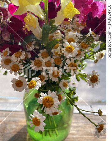 A beautiful bouquet of small white chamomile flowers with golden yellow centers arranged in a translucent green glass vase on a wooden table. Close-up floral photography, rustic home decor A beautiful bouquet of small white chamomile flowers with golden yellow centers arranged in a translucent green glass vase on a wooden table. Close-up floral photography, rustic home decor 143824710