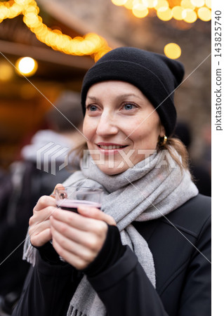 Smiling woman enjoy drink warm mulled wine at Christmas market, wearing black knit hat and gray scarf. Golden festive lights create cozy winter atmosphere during cheerful holiday outing in Germany Smiling woman enjoy drink warm mulled wine at Christmas market, wearing black knit hat and gray scarf. Golden festive lights create cozy winter atmosphere during cheerful holiday outing in Germany 143825740