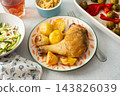 A baked chicken leg rests on a plate with potatoes. Fresh salad, roasted Brussels sprouts, and chickpea bread are arranged on the table. It looks like a wholesome meal 143826039