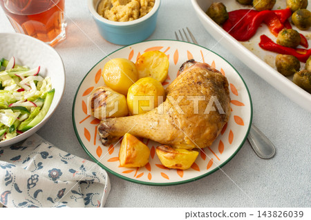 A baked chicken leg rests on a plate with potatoes. Fresh salad, roasted Brussels sprouts, and chickpea bread are arranged on the table. It looks like a wholesome meal 143826039