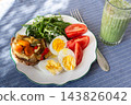 At a breakfast table, a plate holds boiled egg, arugula, tomato, and bread topped with roasted vegetables, with hummus served in a bowl 143826042