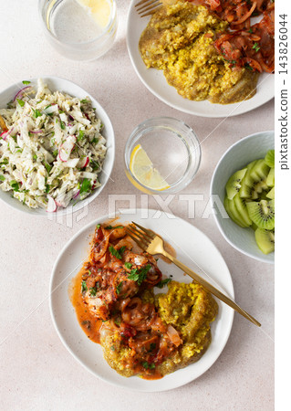Plates hold amaranth, millet, and quinoa porridge served with baked white hake in tomato sauce. Fresh salad is beside the porridge. A bowl of kiwi sits nearby for dessert Plates hold amaranth, millet, and quinoa porridge served with baked white hake in tomato sauce. Fresh salad is beside the porridge. A bowl of kiwi sits nearby for dessert 143826044