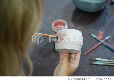 Woman painting pink glaze on handmade ceramic mug with brush in pottery workshop. Pottery decoration represents creativity and learning a craft as a hobby 143826334