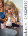 Young woman applying glaze to handmade ceramic mug with brush in pottery workshop. Pottery glazing process represents creativity and artisan craftsmanship 143826340