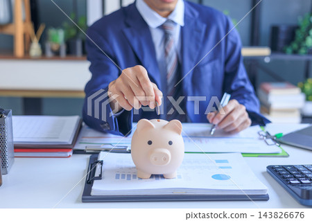 Businessman putting coin into piggy bank on office desk. Concept of saving money. Businessman putting coin into piggy bank on office desk. Concept of saving money. 143826676