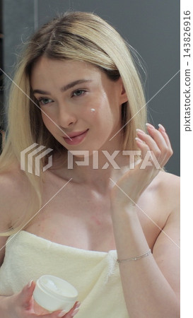 Happy young woman apply skincare cream on face look in bathroom mirror. Facial cream, healthy skin care treatment concept. Happy young woman apply skincare cream on face look in bathroom mirror. Facial cream, healthy skin care treatment concept. 143826916