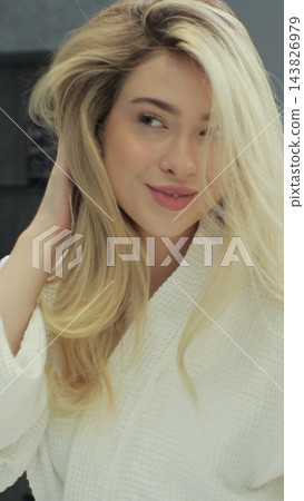 Woman looks into bathroom mirror touches her blond hair. Getting ready in morning, enjoying everyday routine. Wellness and organic skincare. Woman looks into bathroom mirror touches her blond hair. Getting ready in morning, enjoying everyday routine. Wellness and organic skincare. 143826979