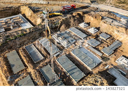Concrete foundation sections and trenches cover a building site, with an excavator and a red truck near the upper edge during daytime Concrete foundation sections and trenches cover a building site, with an excavator and a red truck near the upper edge during daytime 143827321