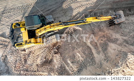 An excavator at a construction site reaches across churned ground with its boom and bucket while tracked treads mark the sandy surface 143827322
