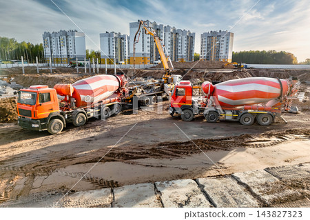 Two concrete mixer trucks stand beside an excavator at a construction site in front of apartment buildings during the day 143827323