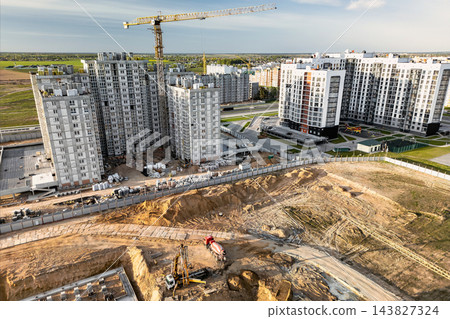 Apartment buildings and a tower crane stand beside a large excavation and dirt roads in a residential district, viewed from above during the day Apartment buildings and a tower crane stand beside a large excavation and dirt roads in a residential district, viewed from above during the day 143827324