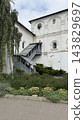 Historic white brick monastery with exterior stairway, garden flowers, and leafy birch tree under cloudy sky 143829697