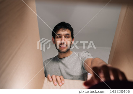 Young man unpacking cardboard box at home, eagerly looking inside receiving new online shopping order, experiencing curiosity and satisfaction. Young man unpacking cardboard box at home, eagerly looking inside receiving new online shopping order, experiencing curiosity and satisfaction. 143830406