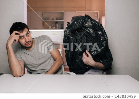 Humorous scene of puzzled young man looking at camera anonymous person sitting nearby with black trash bag over their head. Absurd concept concept of confusion, strange relationships, hiding. 143830459