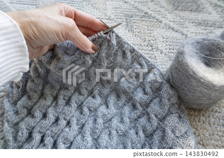 Hands knitting gray cable garment with metal needles. Close-up view of the textured stitches and knitting in progress. Cable sweater on needles. Gray wool knitted sweater. Cozy autumn hobby. Hands knitting gray cable garment with metal needles. Close-up view of the textured stitches and knitting in progress. Cable sweater on needles. Gray wool knitted sweater. Cozy autumn hobby. 143830492