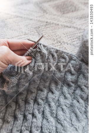 Hands knitting gray cable garment with metal needles. Close-up view of the textured stitches and knitting in progress. Cable sweater on needles. Gray wool knitted sweater. Cozy autumn hobby. Hands knitting gray cable garment with metal needles. Close-up view of the textured stitches and knitting in progress. Cable sweater on needles. Gray wool knitted sweater. Cozy autumn hobby. 143830493