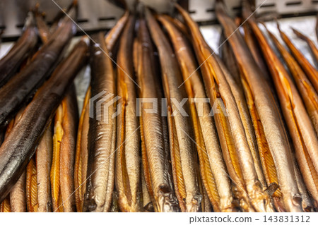 Rows of golden smoked eels hang from metal rack at traditional market, showing glossy skin and rich texture. Close-up seafood display highlights artisanal smoking, preservation and regional cuisine Rows of golden smoked eels hang from metal rack at traditional market, showing glossy skin and rich texture. Close-up seafood display highlights artisanal smoking, preservation and regional cuisine 143831312