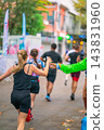 Female Runner Receiving Water from Volunteer, outdoor running. Female runner reaches for water bottle offered by volunteer. Volunteer handing water to marathon runner, hydration station 143831960