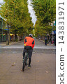 Cyclist in City Street. Cyclist in orange jacket and helmet waits with bicycle on pedestrian street lined with green trees. Cyclist faces road crossing, outdoor sporting activity in urban surroundings 143831971