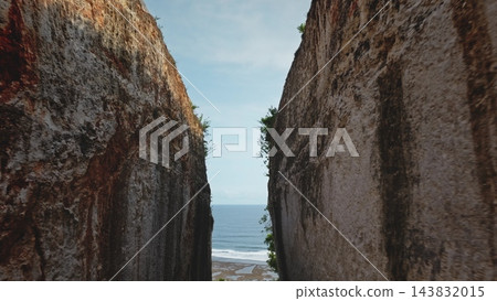 Towering limestone cliffs framing glimpse of tranquil Indian Ocean and sand beach below under bright blue sky. Pathway road winding through a deep limestone canyon in Bali, Indonesia. Drone flight Towering limestone cliffs framing glimpse of tranquil Indian Ocean and sand beach below under bright blue sky. Pathway road winding through a deep limestone canyon in Bali, Indonesia. Drone flight 143832015