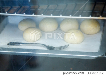 Fresh dough balls rising on parchment lined baking tray inside warm oven with metal fork in foreground, soft steam haze, home baking preparation, simple kitchen food process Fresh dough balls rising on parchment lined baking tray inside warm oven with metal fork in foreground, soft steam haze, home baking preparation, simple kitchen food process 143832147