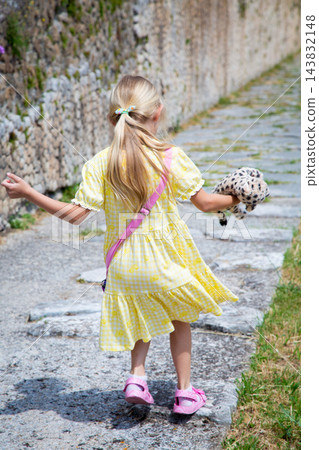 Young girl walking along stone path beside textured wall, holding plush toy and wearing yellow dress, pink sandals, and crossbody bag, seen from behind in soft daylight 143832148
