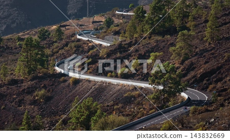 Tenerife, Canary Islands: Aerial view capturing cars riding winding road through Tenerife's volcanic terrain, with lush pine trees contrasting dark rock. Static shot Tenerife, Canary Islands: Aerial view capturing cars riding winding road through Tenerife's volcanic terrain, with lush pine trees contrasting dark rock. Static shot 143832669