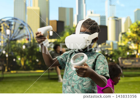 African child with virtual reality headset standing on picnic blanket and holding controllers as parents relax beside food and basket in sunny urban park. VR interactive technology. African child with virtual reality headset standing on picnic blanket and holding controllers as parents relax beside food and basket in sunny urban park. VR interactive technology. 143833356
