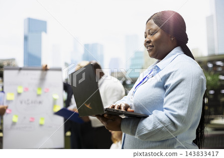 Businesswoman, laptop and typing as professional works while colleagues meet by strategy board. Productivity, digital workflow and coordination support flexible work on outdoor office terrace. 143833417